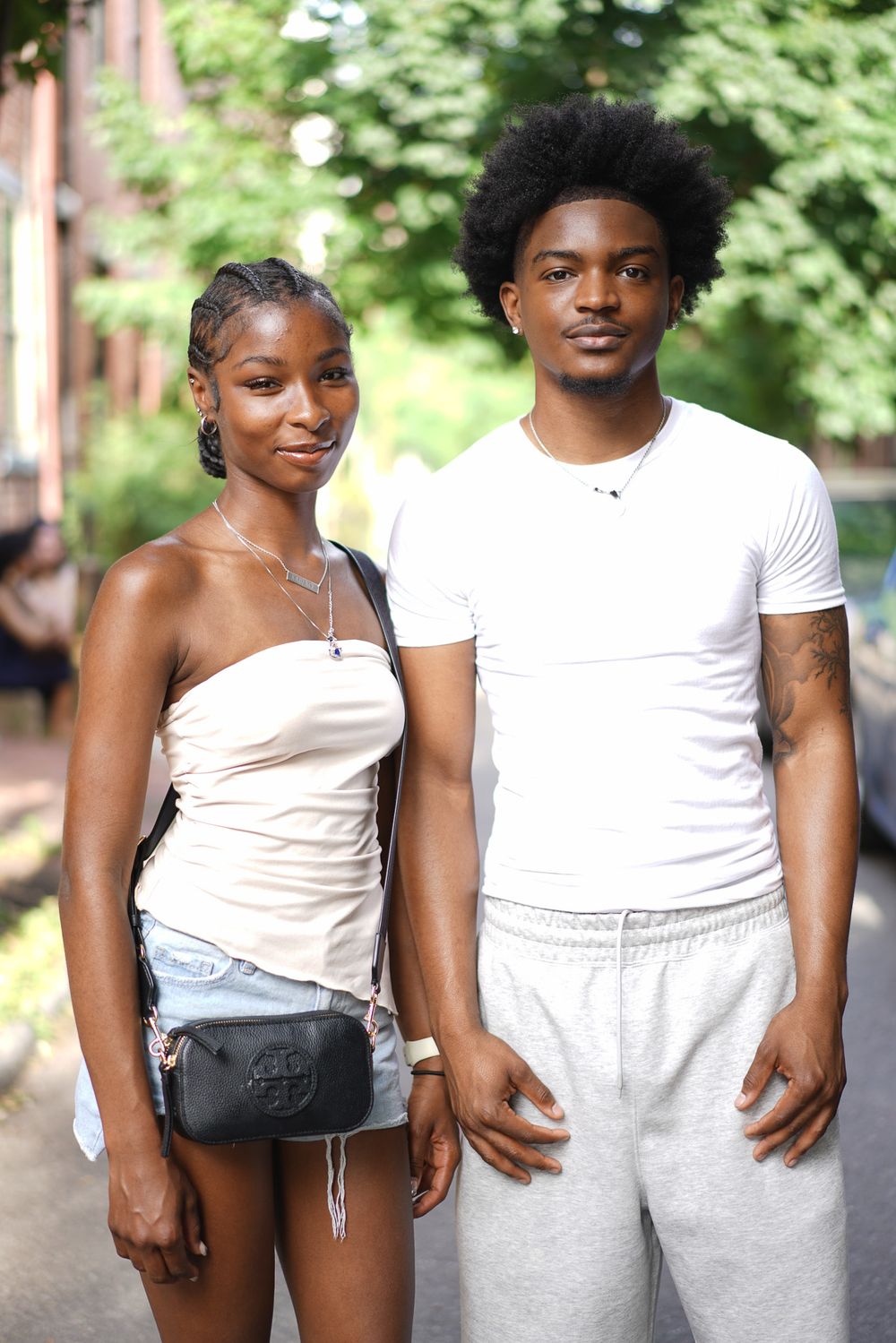Young couple posing outdoors, woman in white tube top with crossbody bag, man in white t-shirt and gray sweatpants.