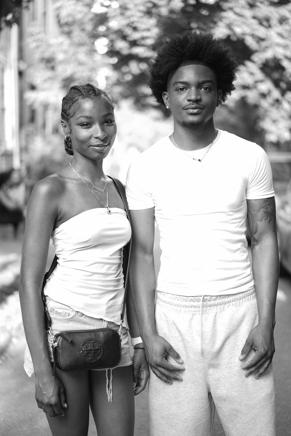 Black and white portrait of a young couple in white outfits posing outdoors on a tree-lined street.