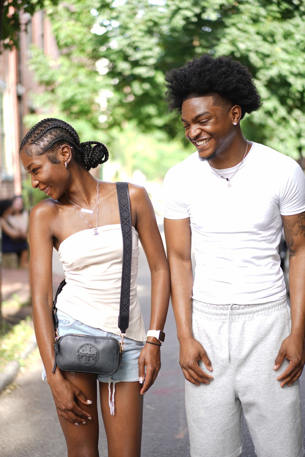 A smiling young couple in casual white outfits walks together on a tree-lined path.