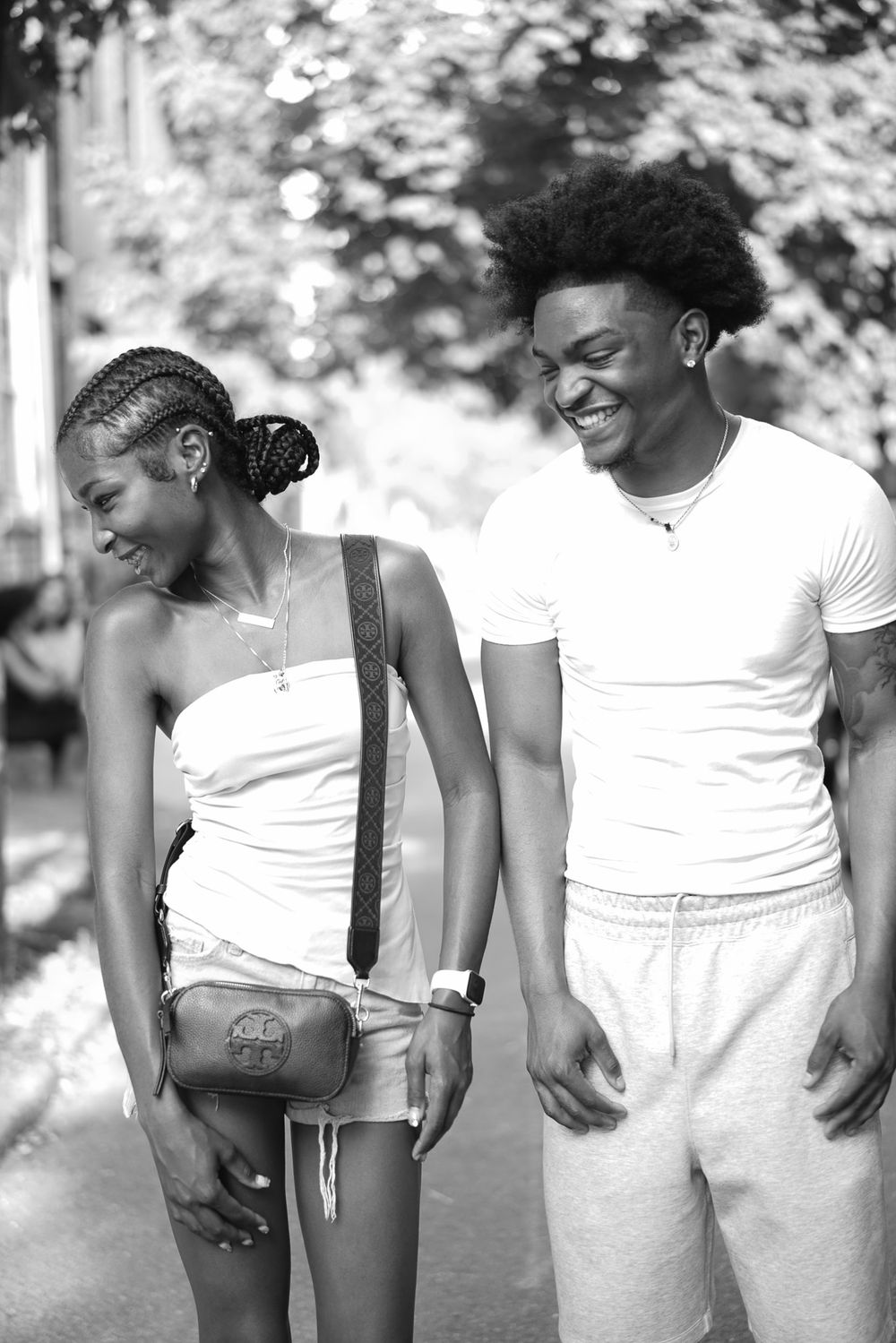 Black and white photo of a young couple laughing together outdoors, wearing white outfits on a sunny day.