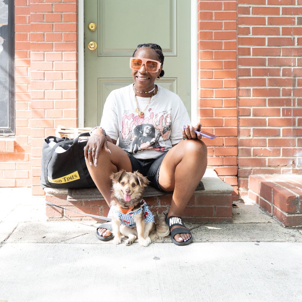 Person with orange sunglasses sits on brick steps with small dog wearing bandana on sunny day.