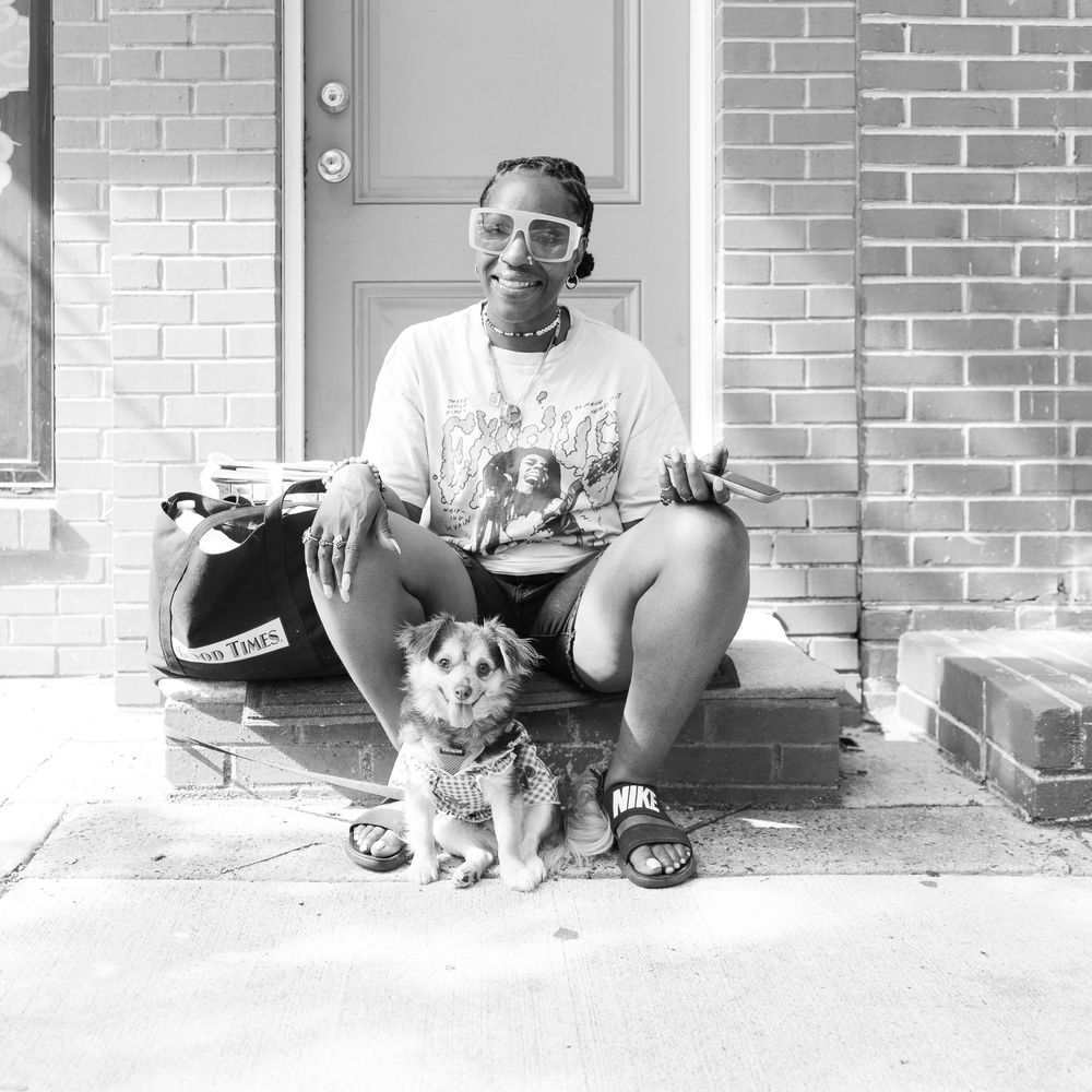 Black and white photo of a person sitting on front steps with a small dog, wearing sunglasses and sandals.