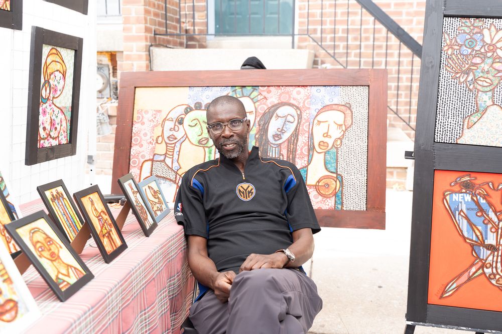 African artist sitting outdoors surrounded by colorful framed paintings at an art exhibition display.