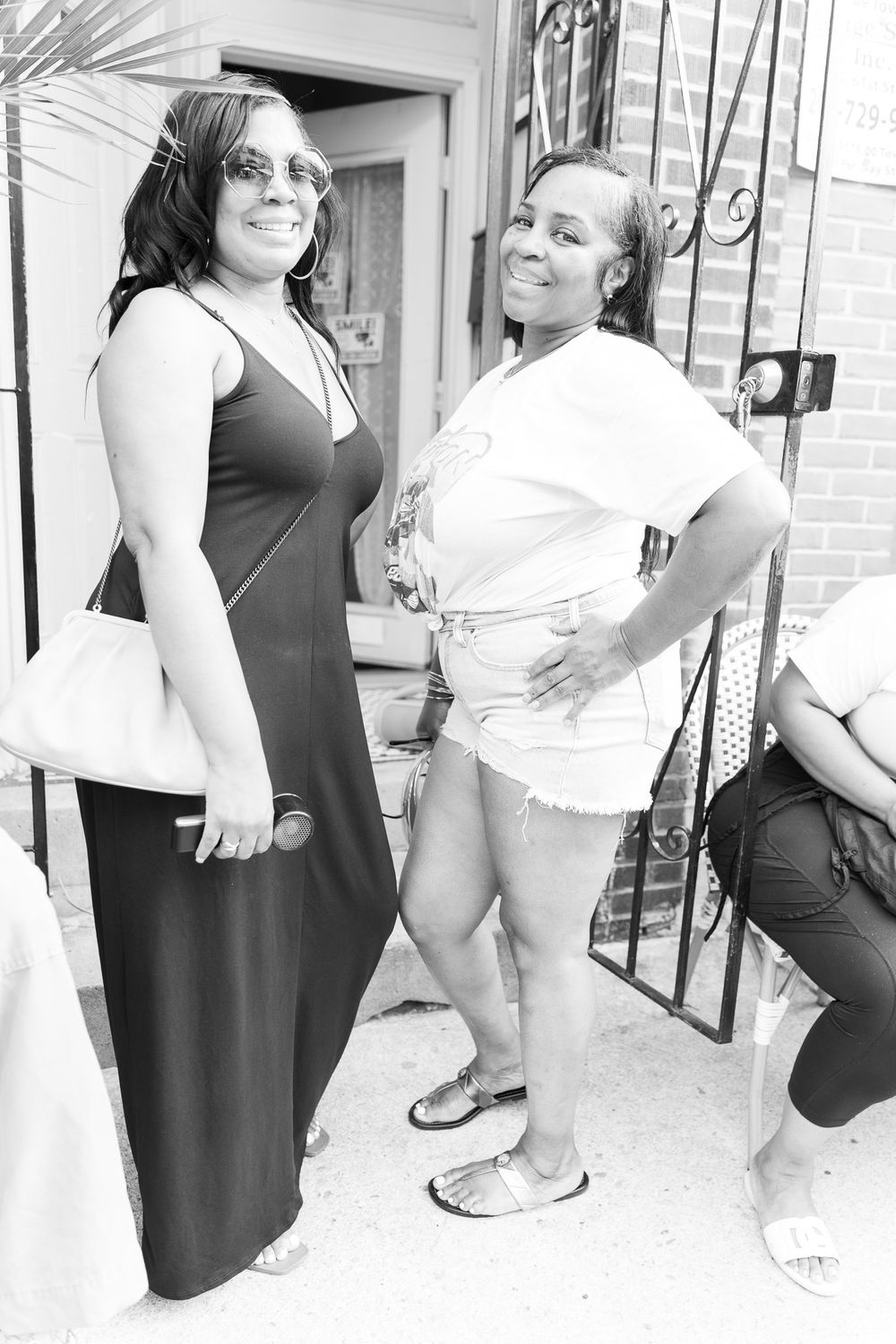 Two women posing outdoors in black and white photo, one in maxi dress with handbag, other in shorts and white top.