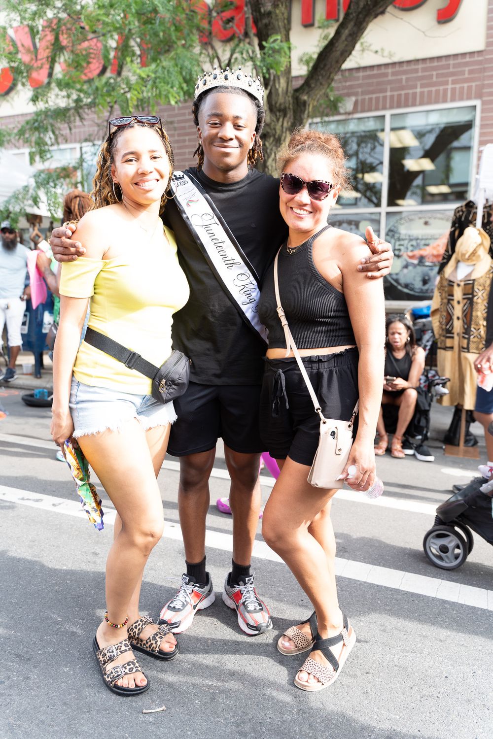 Three friends pose together at an outdoor street festival, with one wearing a pageant sash and crown.