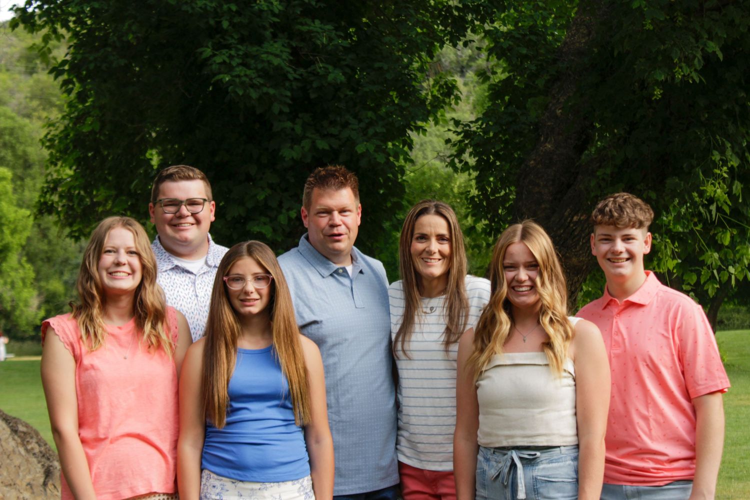 A cheerful family of seven poses outdoors, smiling together against a lush green tree backdrop.