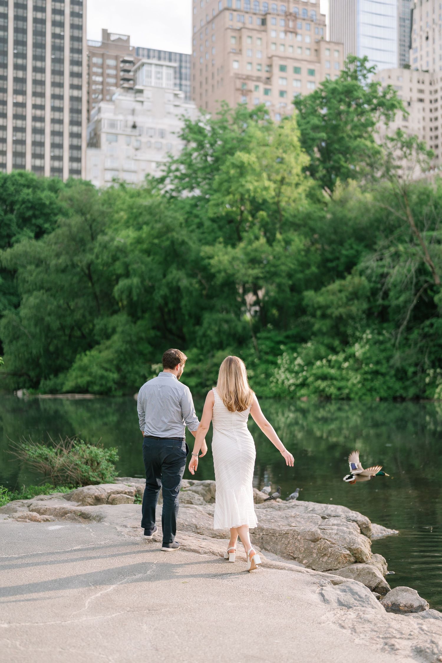 Couple holding hands walking along Central Park pond with NYC skyline and lush green trees in background.
