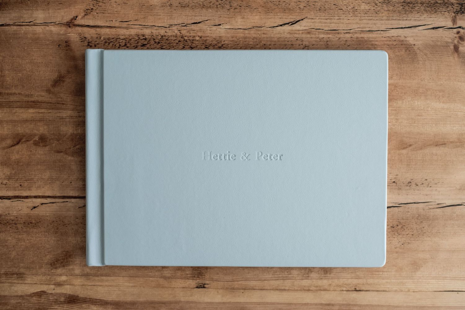 Light blue linen wedding photo album with embossed names on wooden table background.
