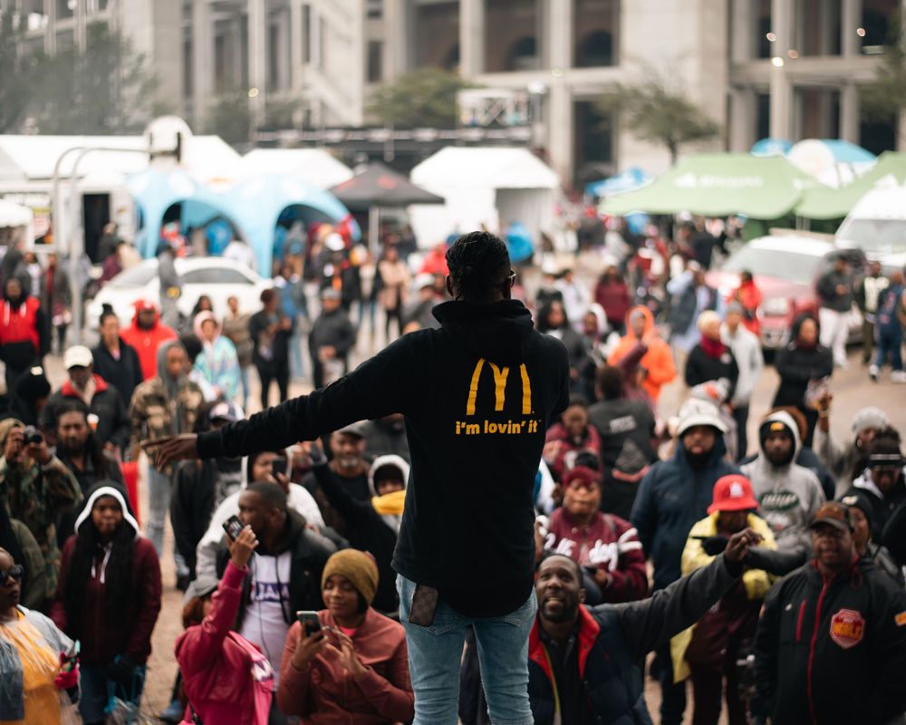 Person in McDonald's hoodie stands before a large crowd at an outdoor urban gathering.