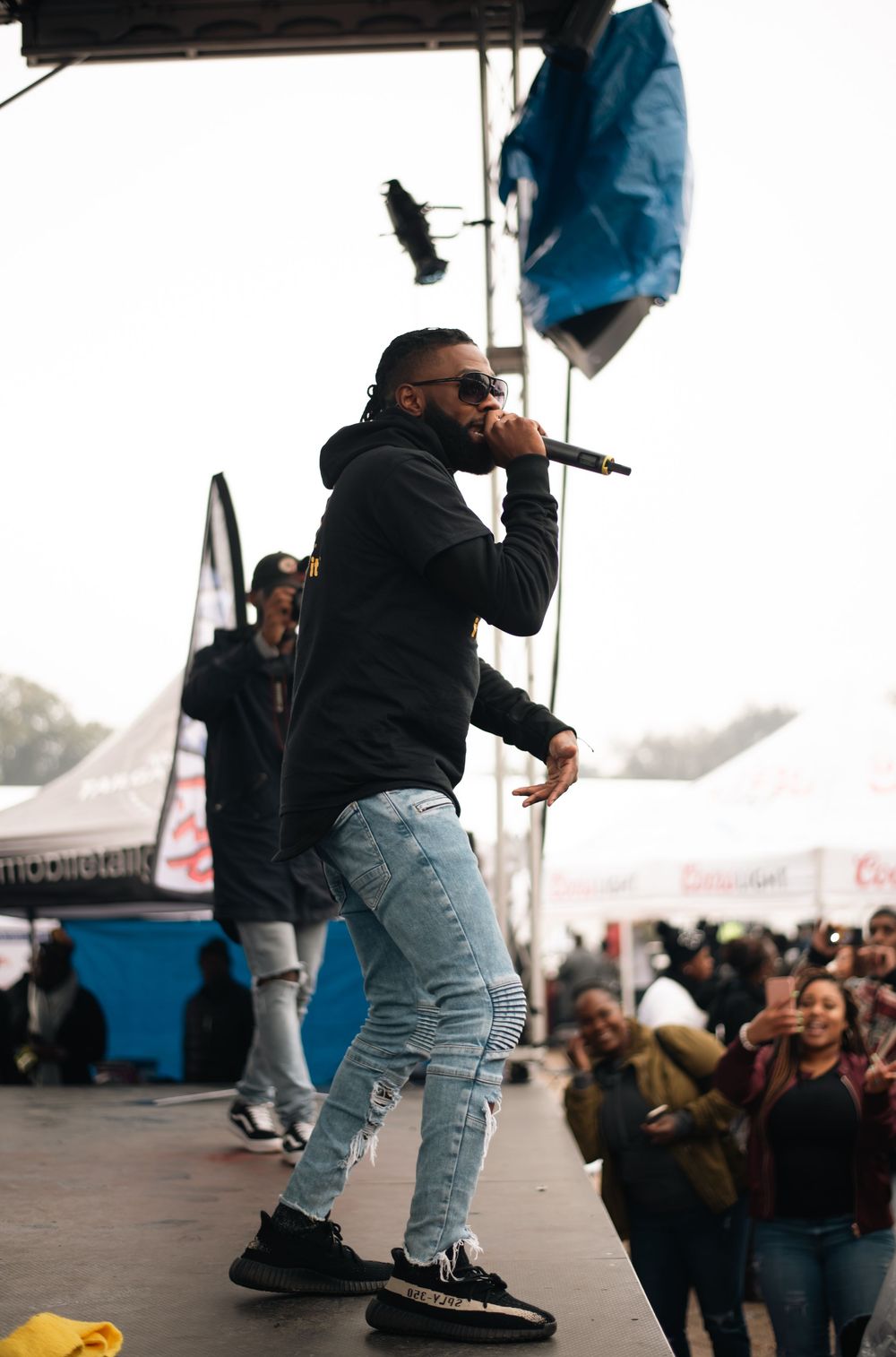 Man performing on stage holding microphone wearing black hoodie and light blue ripped jeans with Yeezy sneakers at outdoor festival.