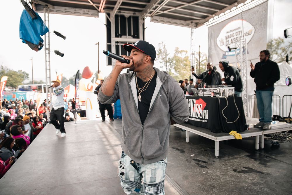 Rapper performing on outdoor stage with microphone, wearing gray hoodie and cap at a music festival.