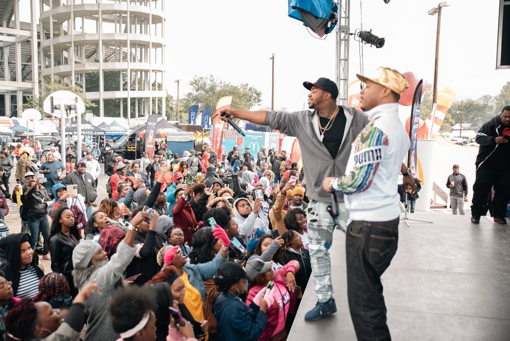 Two performers on stage energize a large outdoor festival crowd, with one extending a microphone toward fans.