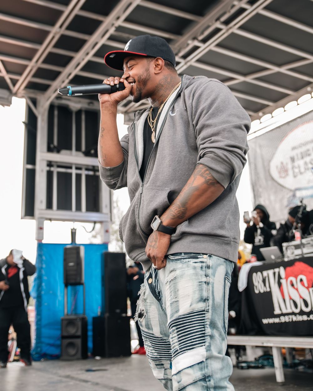 Man in gray hoodie and ripped jeans performs on outdoor stage holding microphone with radio station banner behind him.