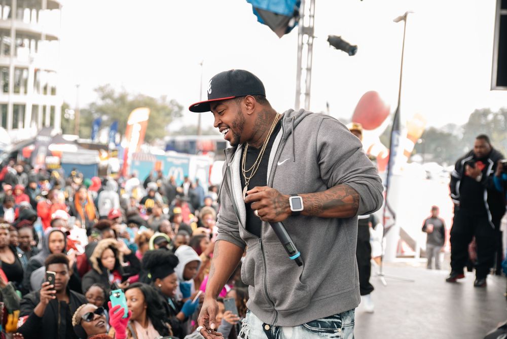 A performer in a gray hoodie and cap smiles on stage, holding a mic before a large outdoor crowd.