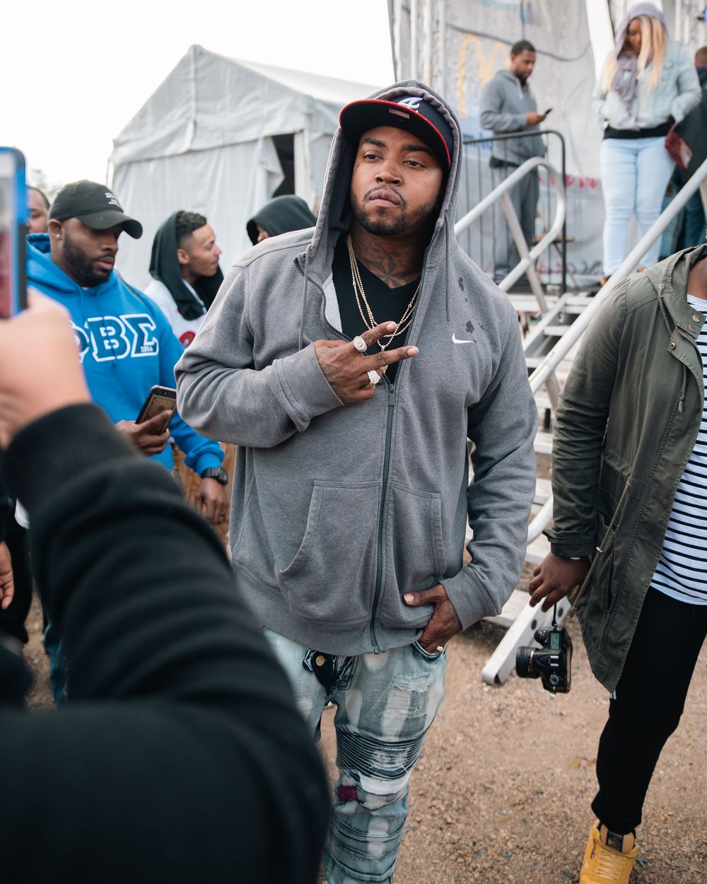 Man in gray Nike hoodie and ripped jeans posing at outdoor event while someone photographs him.