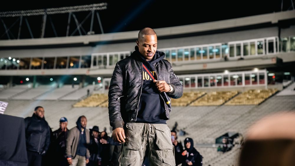 Man in black jacket and cargo pants stands on stadium field at night with crowd in background.