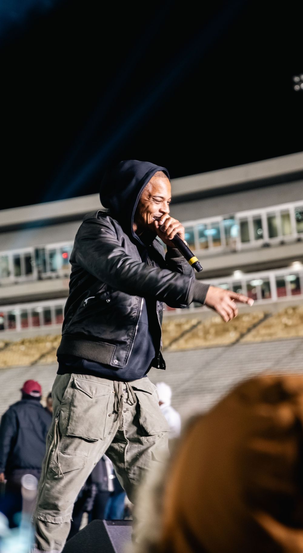 A performer in a hoodie and cargo pants raps into a microphone on an outdoor stage at a live concert event.