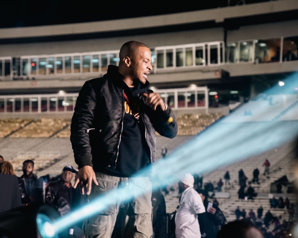 A man performs on stage at a nighttime outdoor concert, holding a microphone with bright stage lights beaming behind him.