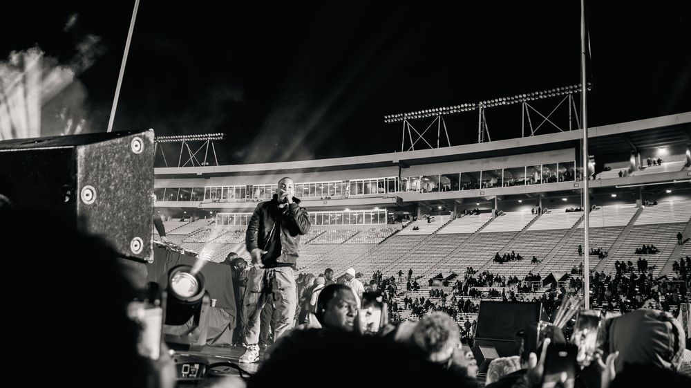 Black and white photo of a performer on stage at a packed outdoor stadium concert at night with dramatic lighting.