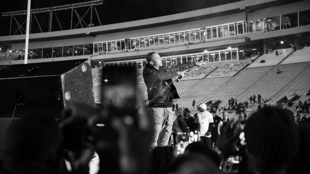 Black and white photo of a speaker addressing a large crowd at a nighttime stadium event.