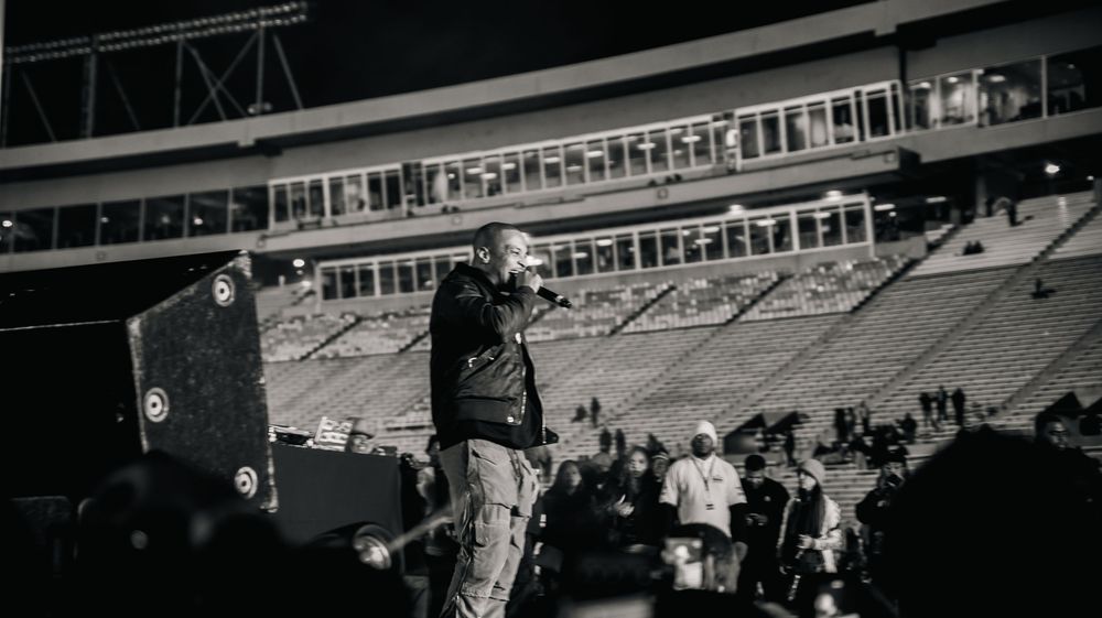 A performer speaks into a microphone on a dark stadium stage at night with empty bleachers in the background.