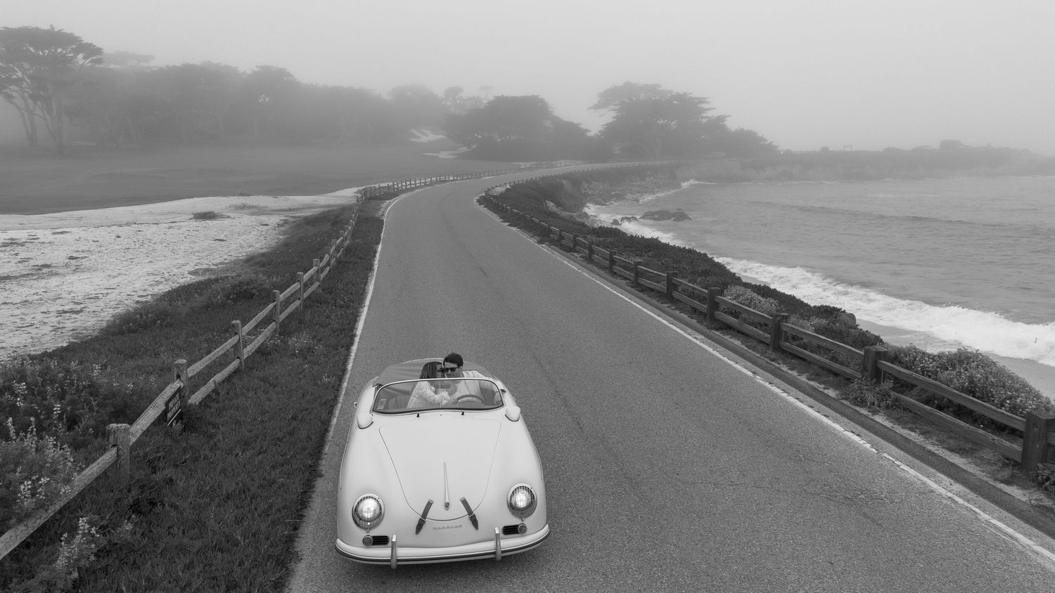 Vintage white Porsche convertible driving along a misty coastal road in a classic black and white photograph.