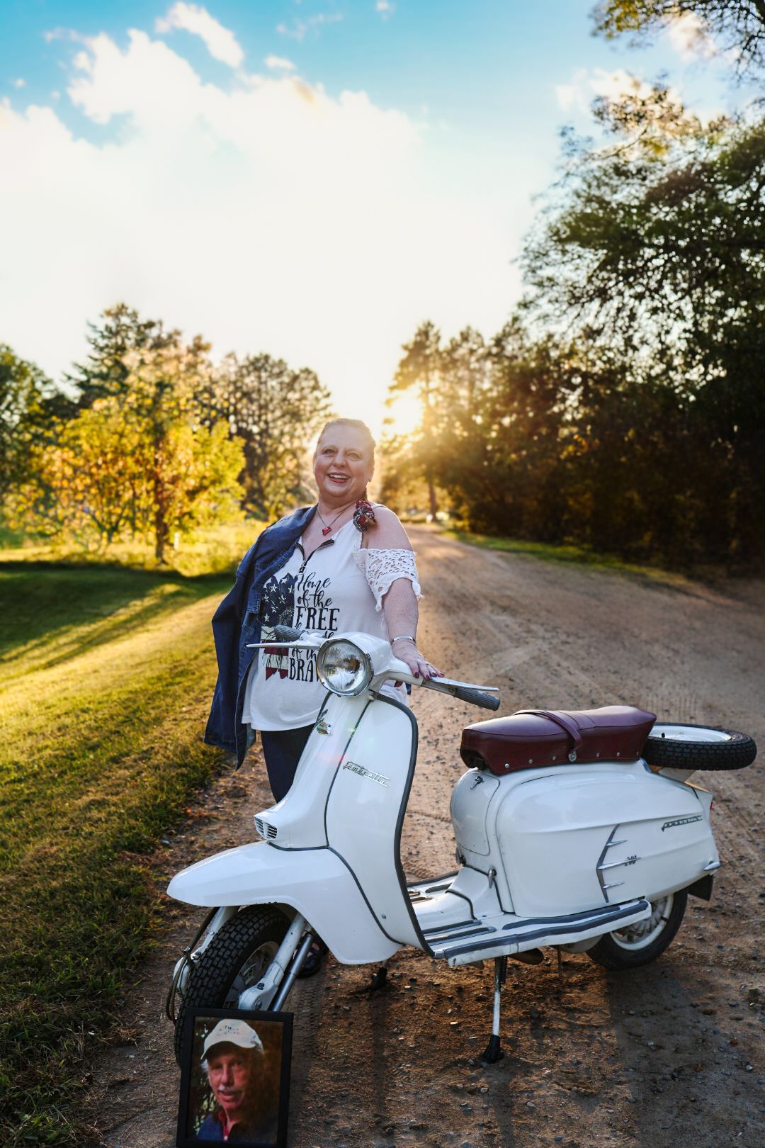 Young woman in white off-shoulder dress poses outdoors at sunset near a lake with autumn trees in background.