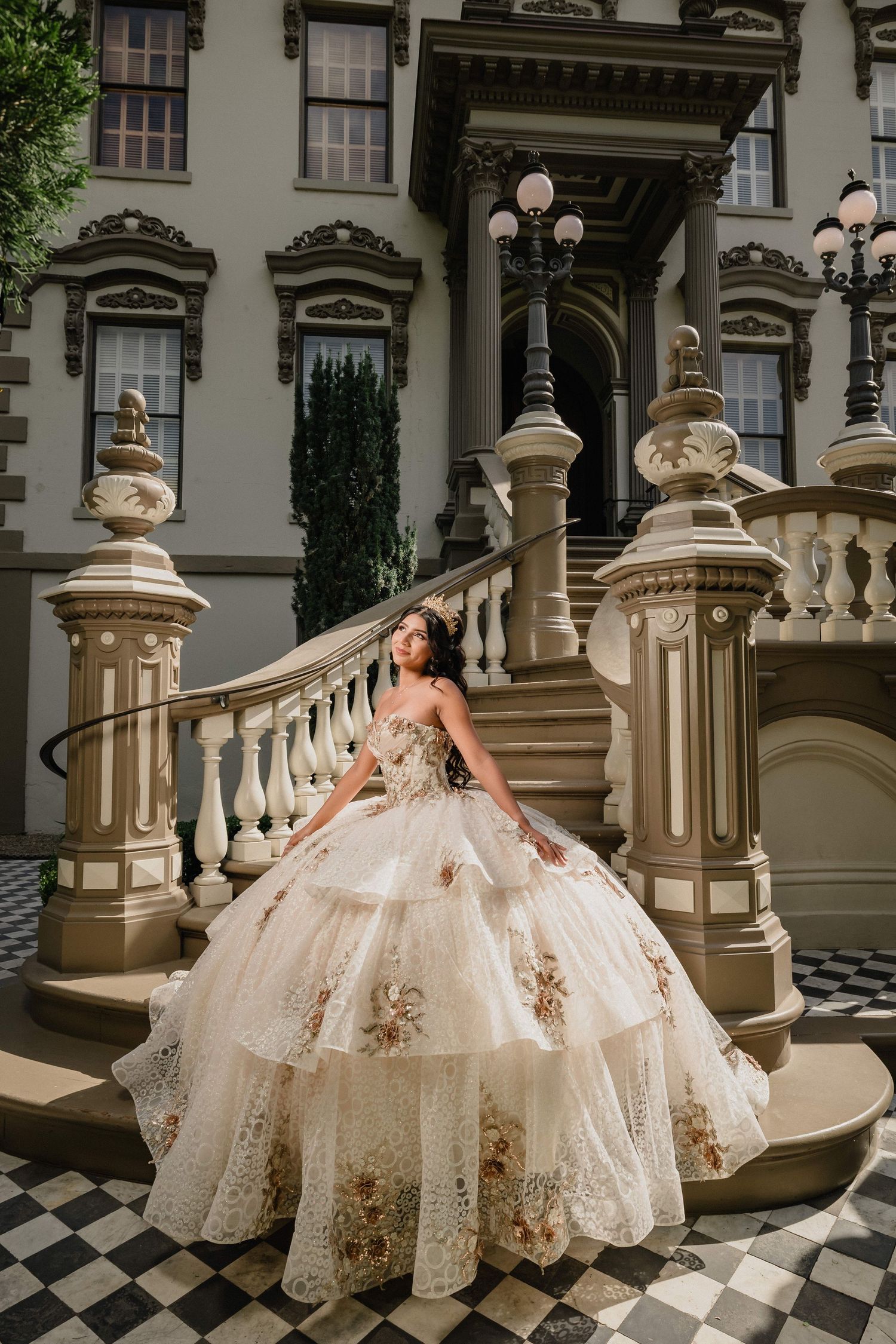 Young woman in an ornate floral ballgown poses on grand staircase of elegant historic mansion.