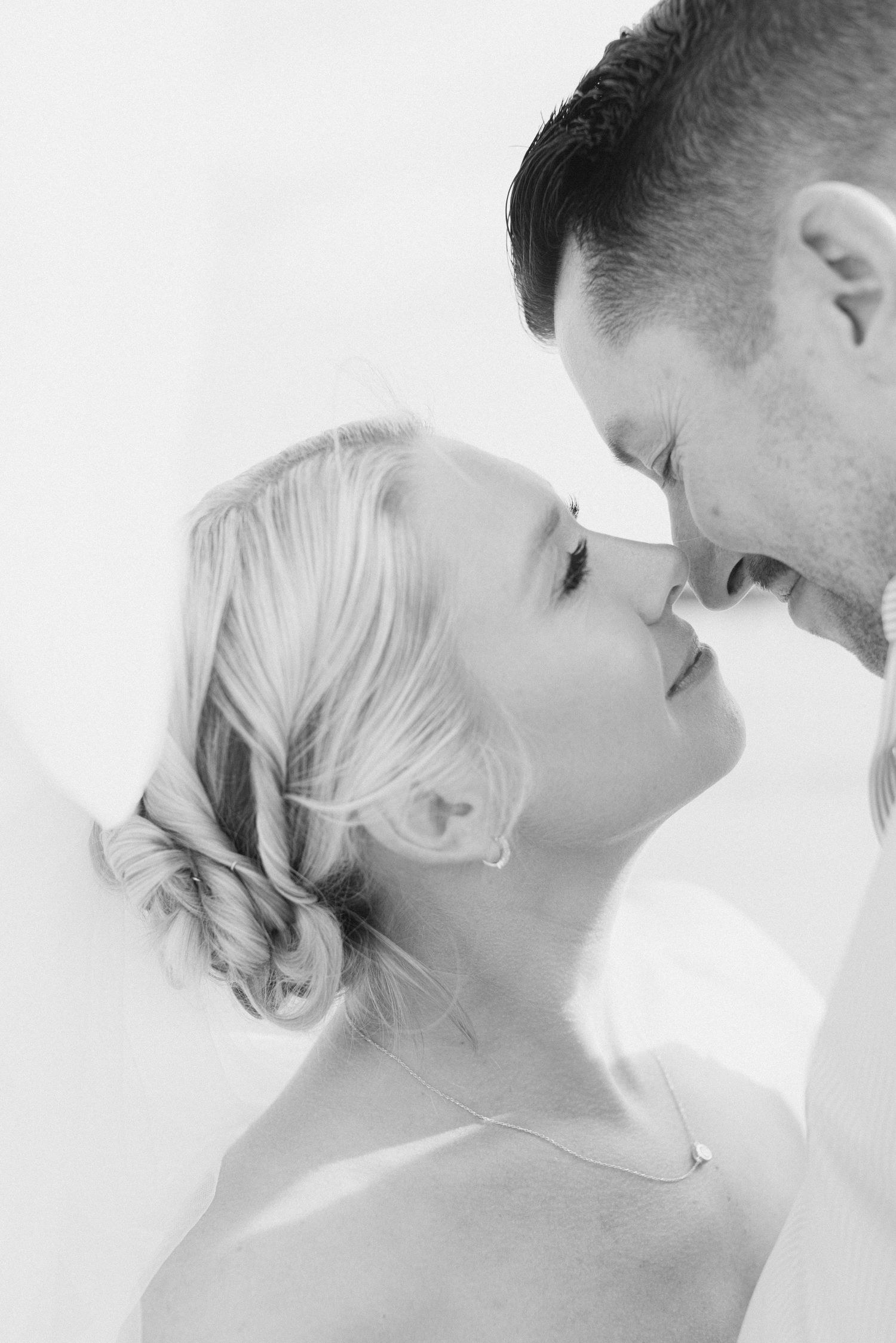 Black and white photo of a couple sharing a tender moment, noses touching, bride with elegant updo and delicate necklace.
