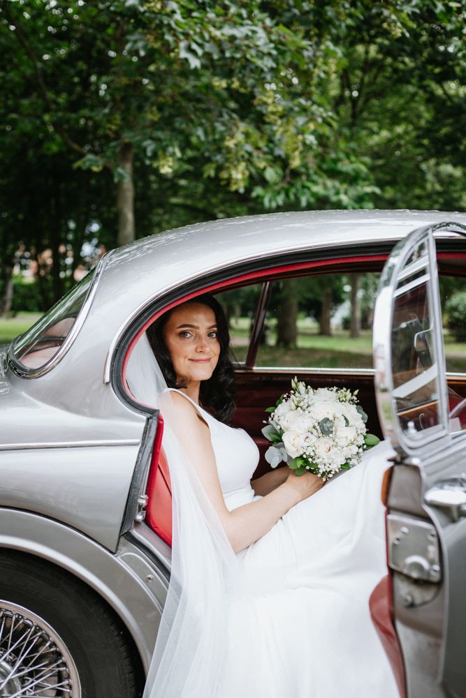 Bride in white gown holding bouquet of white and cream flower seated in vintage silver car with red interior, west bridgford, nottingham