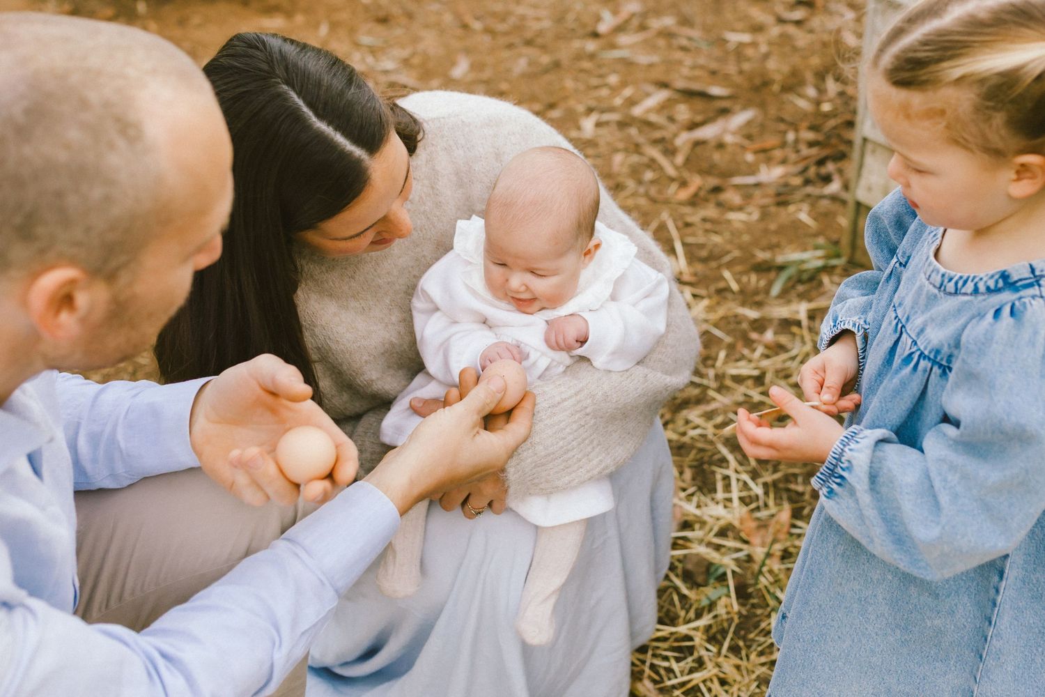Family with baby and toddler girl examining eggs outdoors on hay-covered ground in a warm, natural setting.