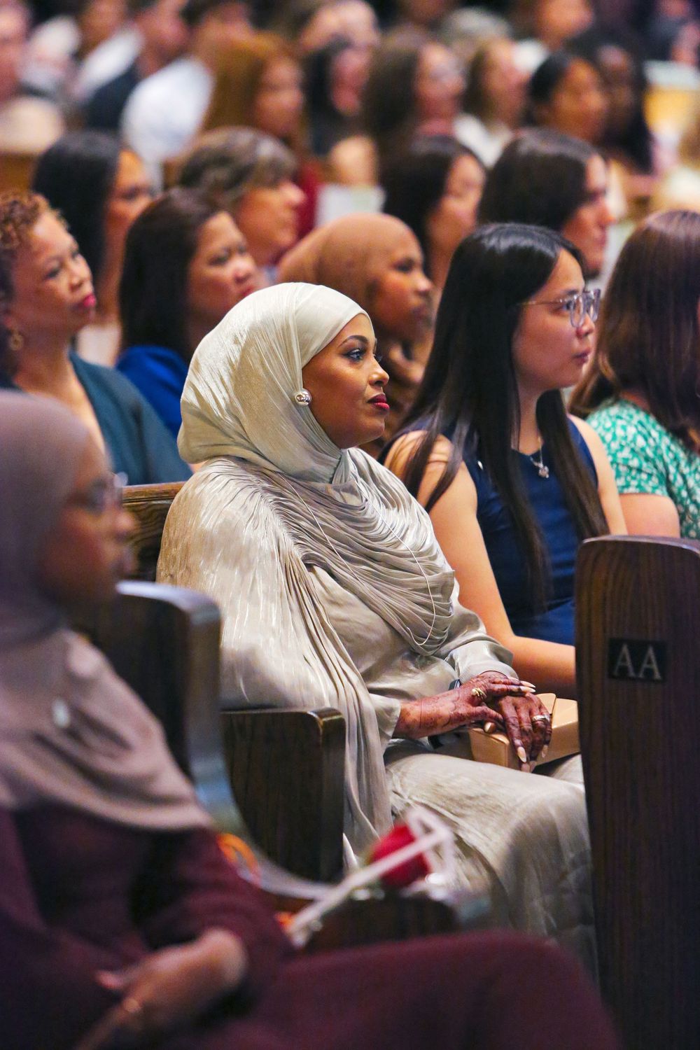Diverse audience seated in auditorium, woman in elegant white hijab prominently featured among attendees at formal event.