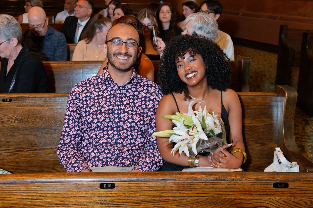 Couple smiling in church pew, man in floral shirt, woman with curly hair holding white lilies bouquet.