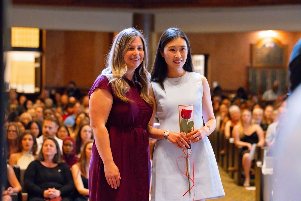Two women smiling together at a formal ceremony, one holding a red rose, with a seated audience in the background.