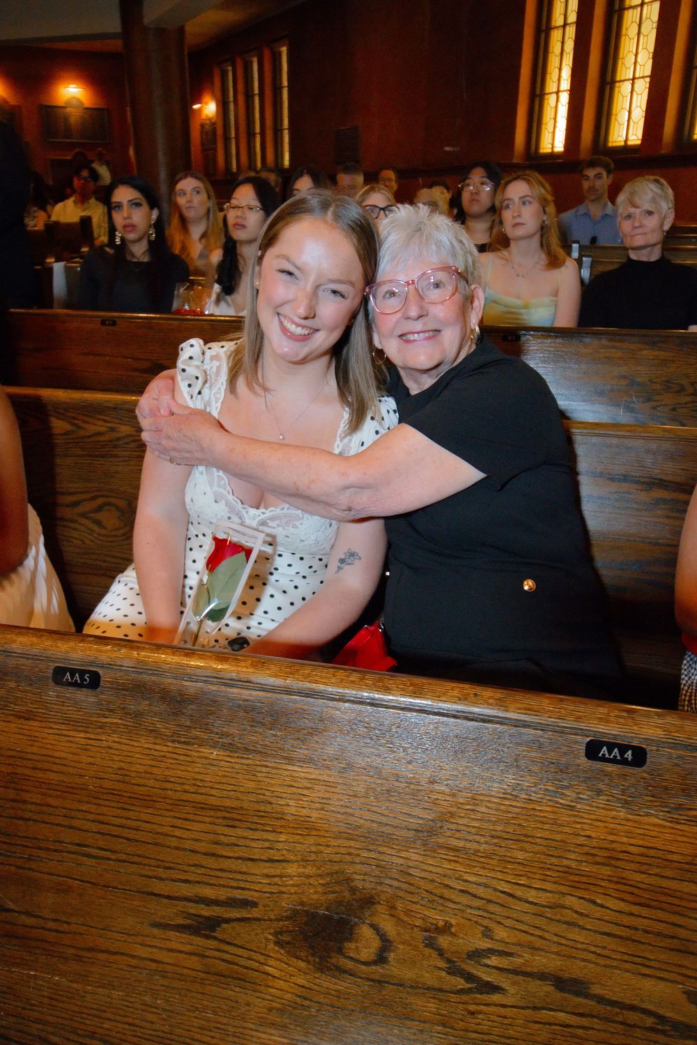 Two women smile and hug while seated in wooden church pews during a formal ceremony.