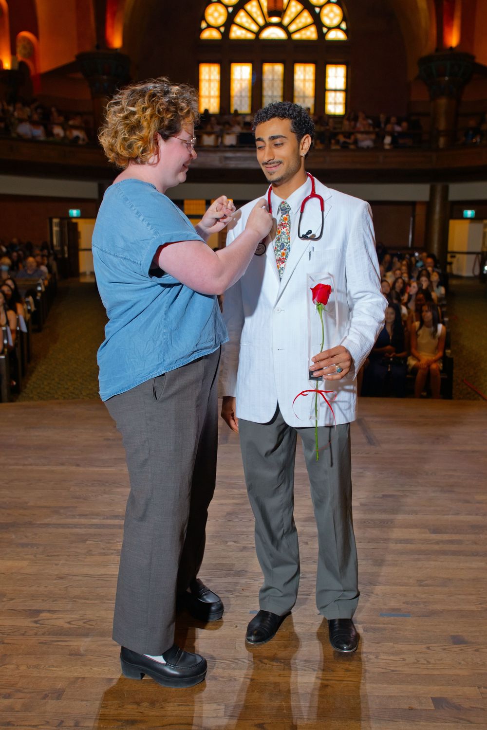 Medical student in white coat holds red rose while receiving pin during white coat ceremony in grand auditorium.