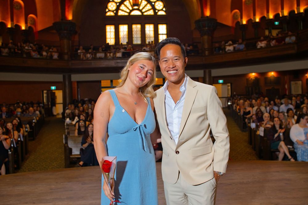 A man and woman smile together in a grand auditorium filled with guests at a formal event.