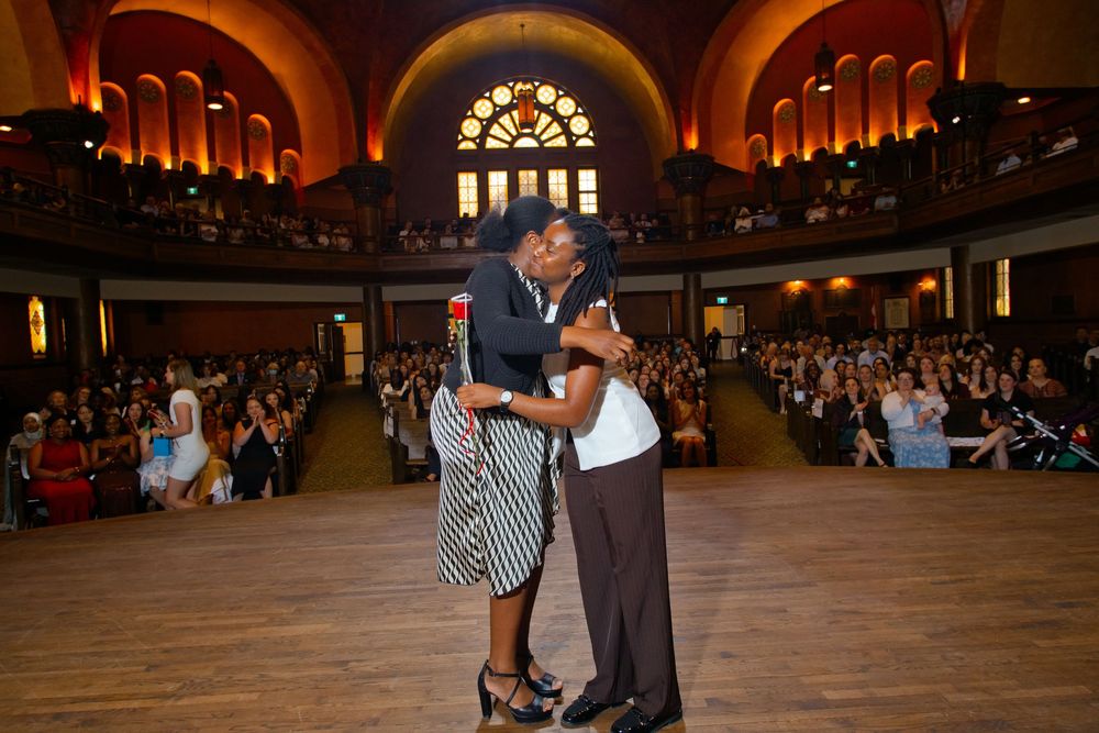 Two people share an embrace while dancing in an elegant arched hall filled with seated audience members.