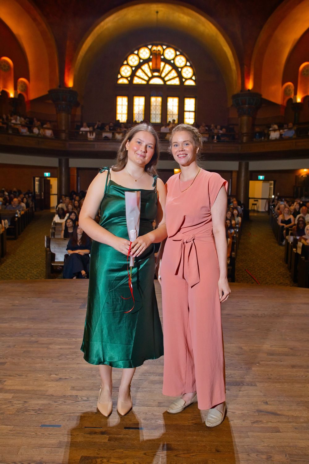 Two women pose together in an elegant hall, one in green satin dress, one in pink jumpsuit, at a graduation ceremony.
