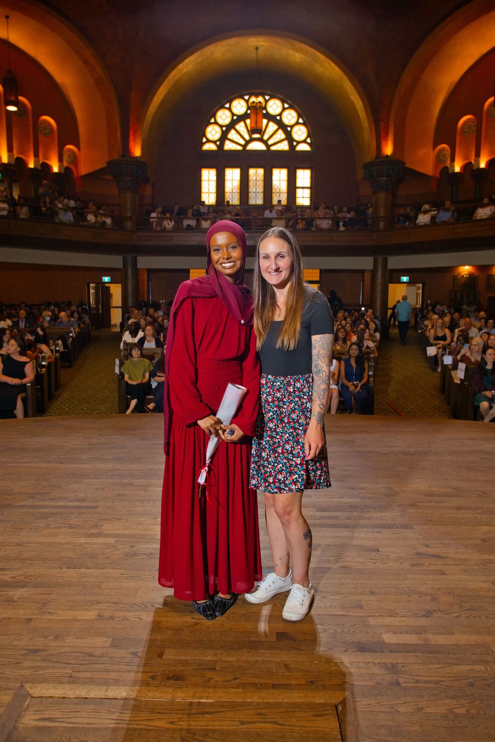 Two women pose together at a graduation ceremony in an ornate hall filled with seated guests.