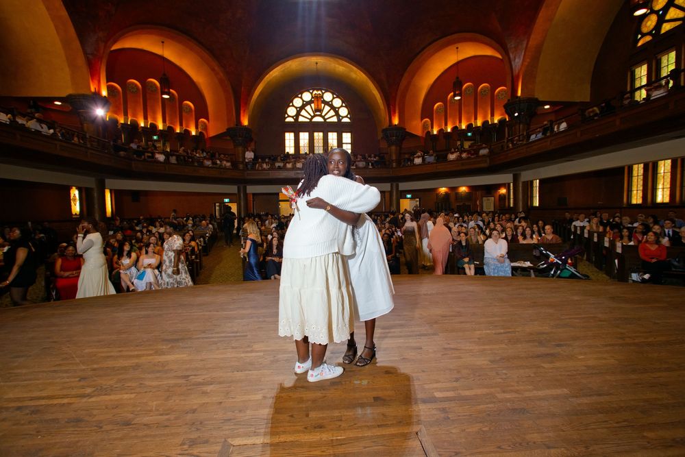 Two women embrace on a dance floor in an elegant grand hall filled with seated guests during a formal event.