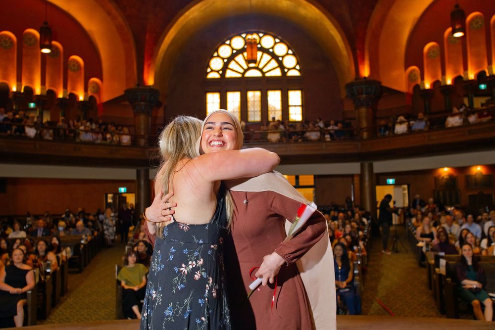 Two women share warm embrace during graduation ceremony in ornate university auditorium with audience.