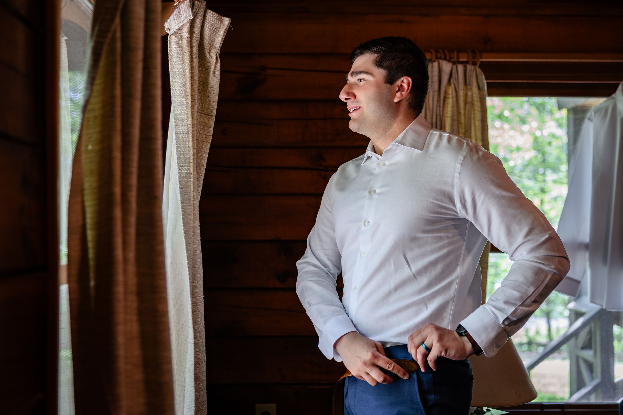 Groom in white dress shirt gazes out cabin window while getting ready on his wedding day.