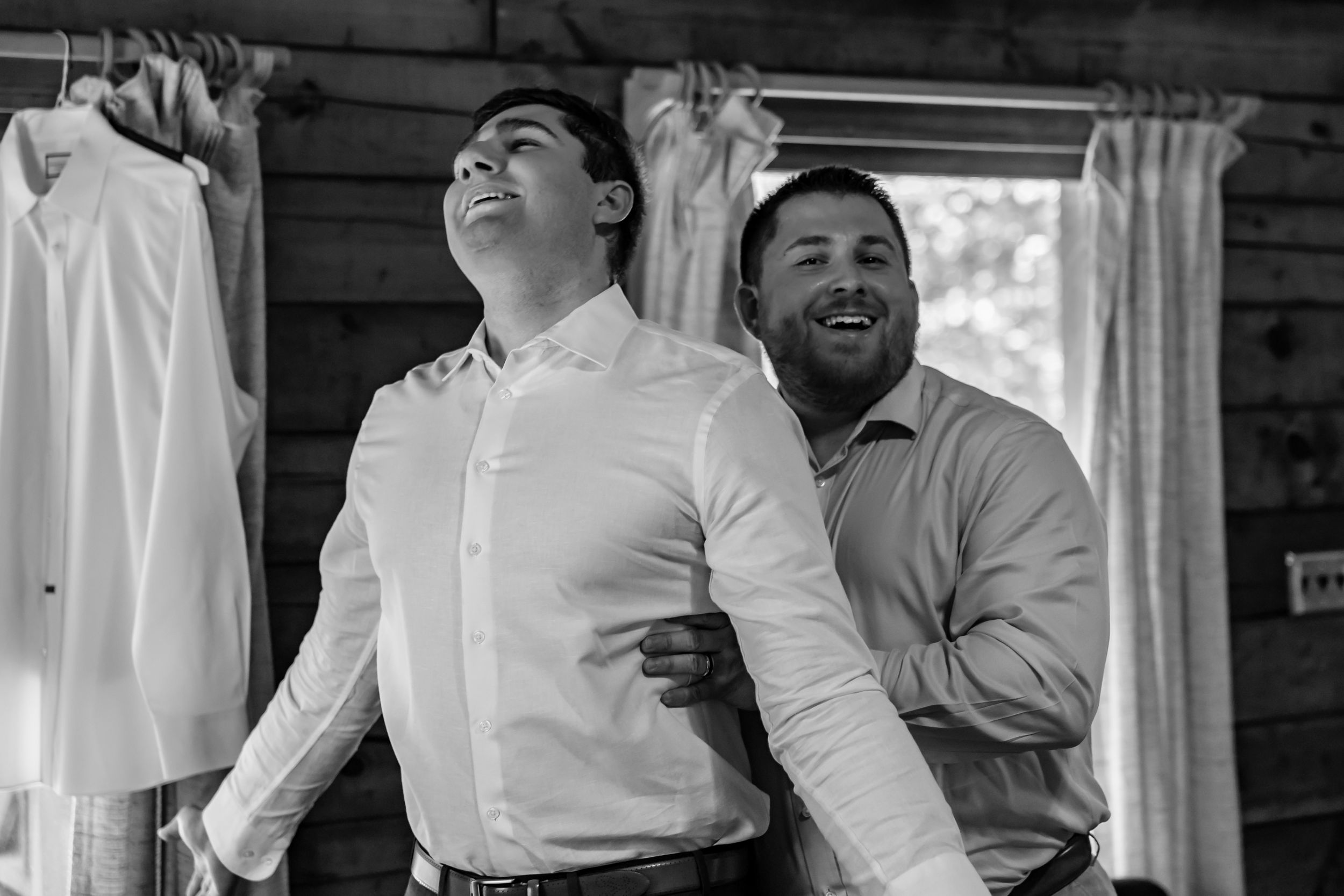 Two men laughing and joking while getting dressed in formal shirts at a rustic wooden cabin wedding preparation.