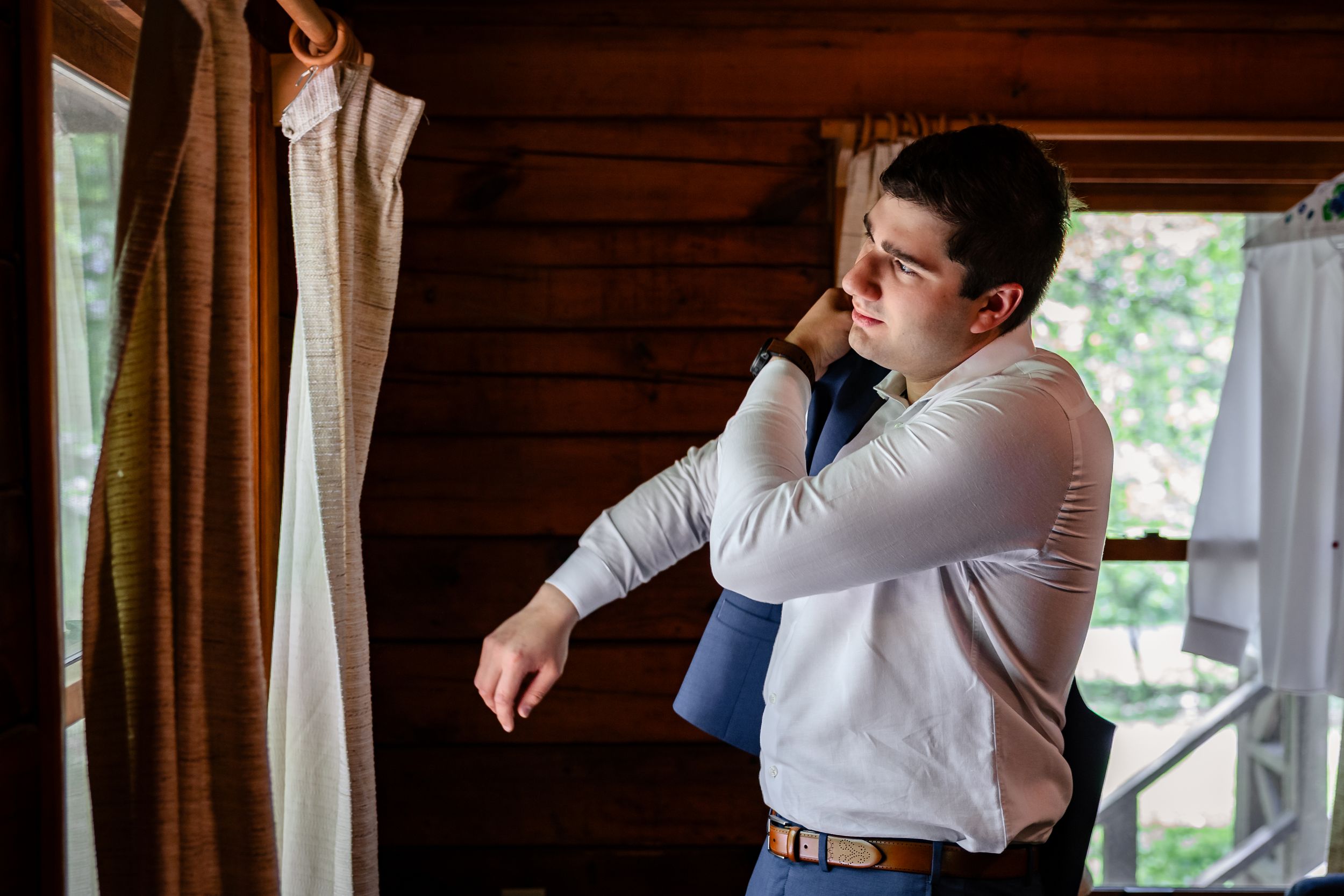 Groom adjusting his tie while getting ready near a window in a rustic wooden cabin before his wedding.