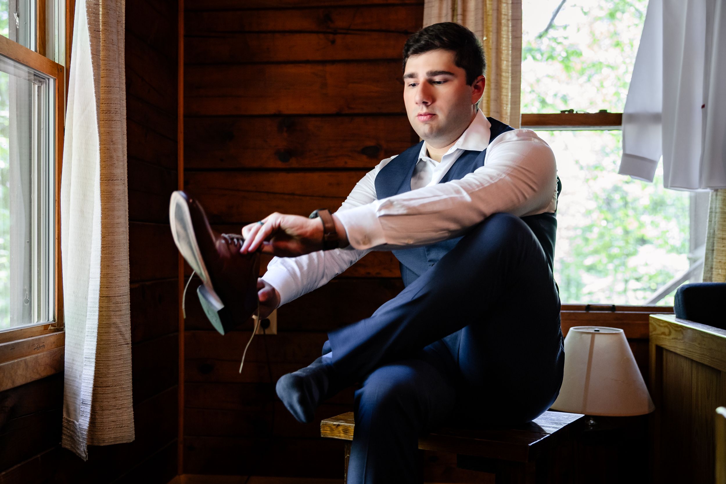 Groom putting on dress shoes while seated by a window in a rustic wooden cabin before his wedding.