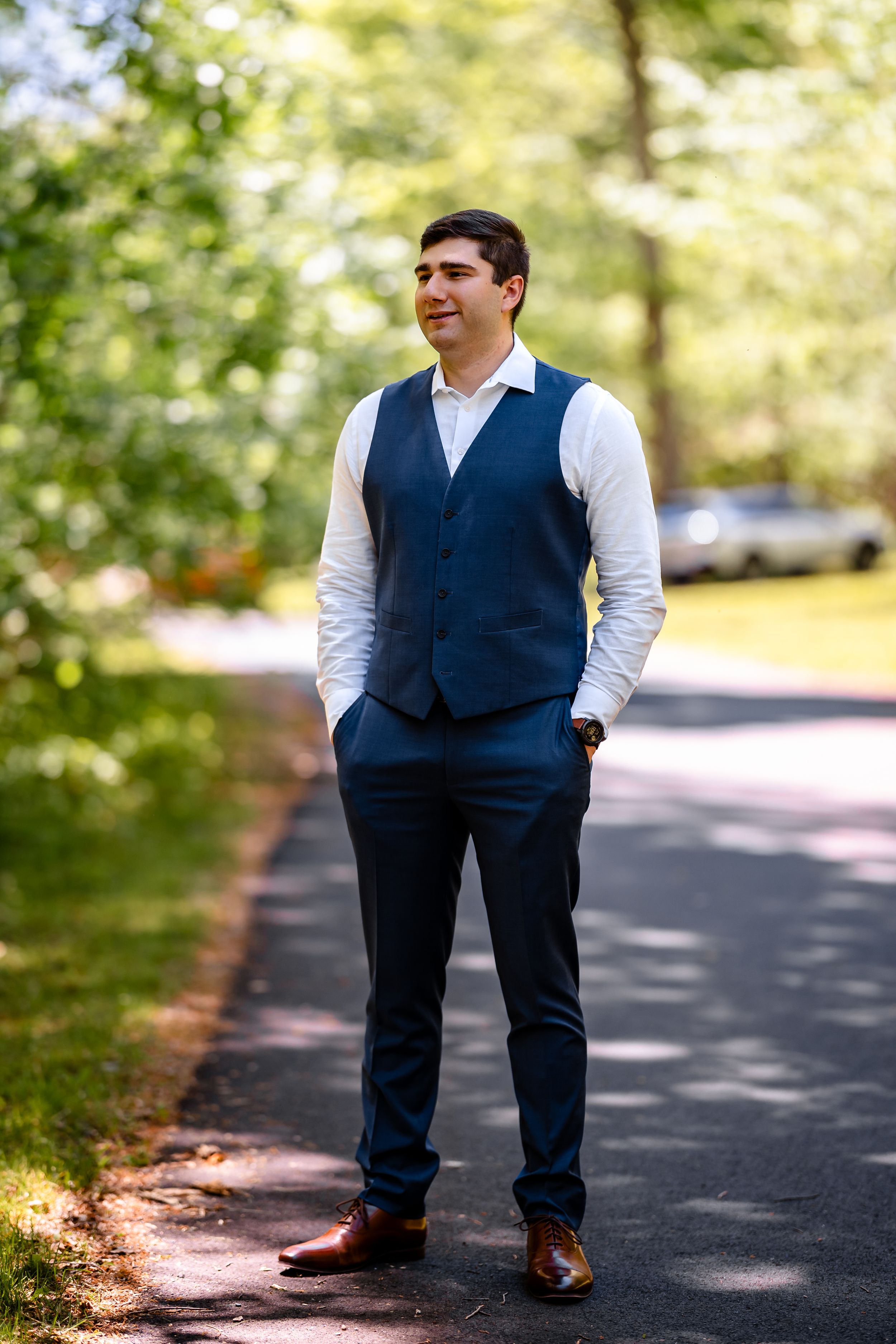 Groom in navy blue vest and trousers standing on sunny outdoor pathway.