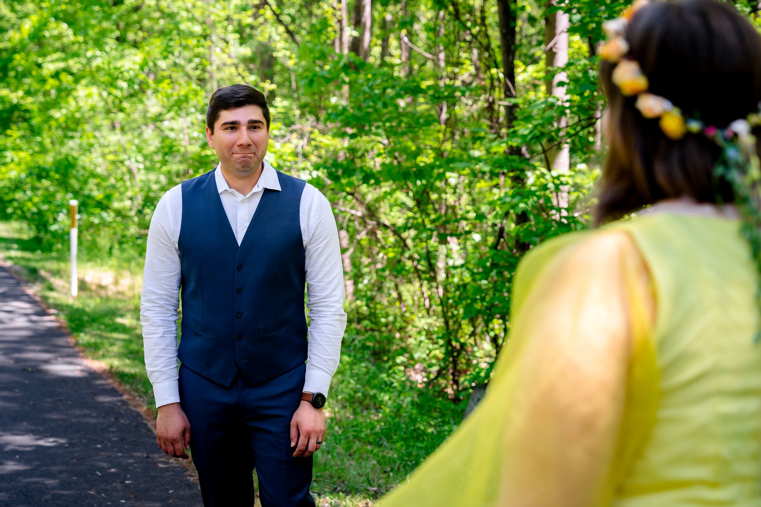 Groom in navy vest smiles at bride in yellow dress on a lush green forest path.