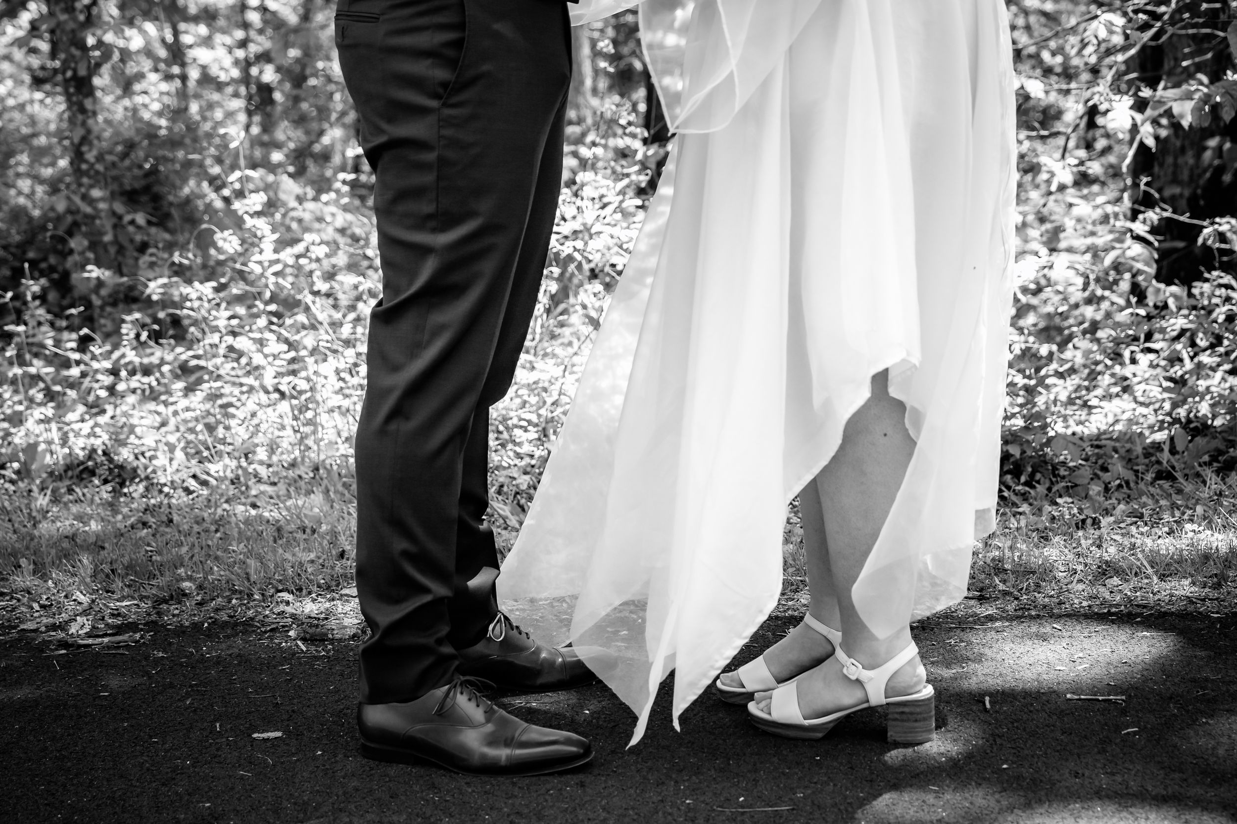 Black and white photo of bride and groom standing close, showing wedding dress hem and dress shoes on pavement.