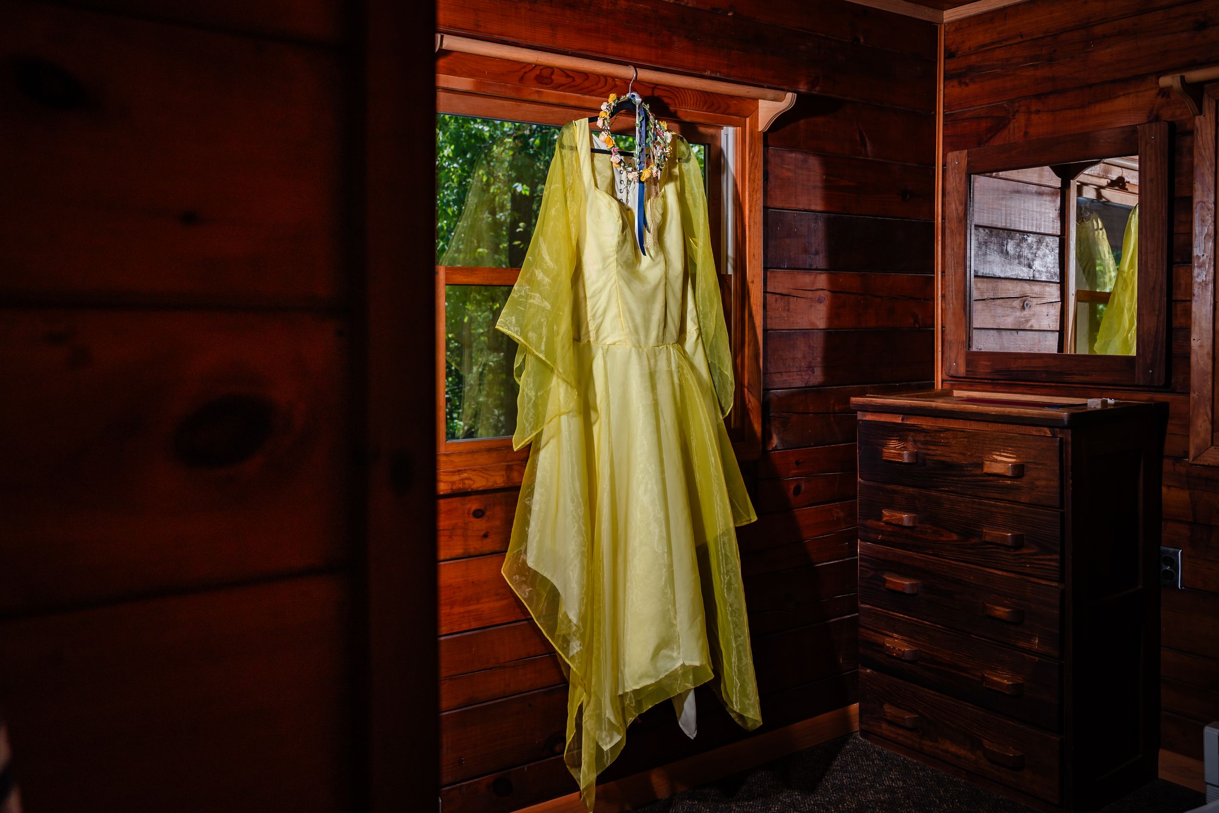 Yellow wedding dress hanging in rustic wooden cabin with floral necklace accessory.